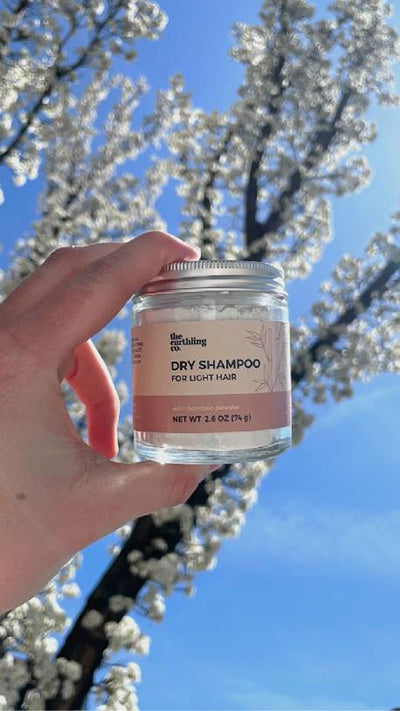 a woman holding a jar of earthling co dry shampoo in front of a tree with blooming flowers